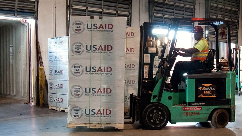 Stacked cardboard cases on a warehouse pallet with a shrink-wrapped corner and a printed delivery manifest taped on top.
