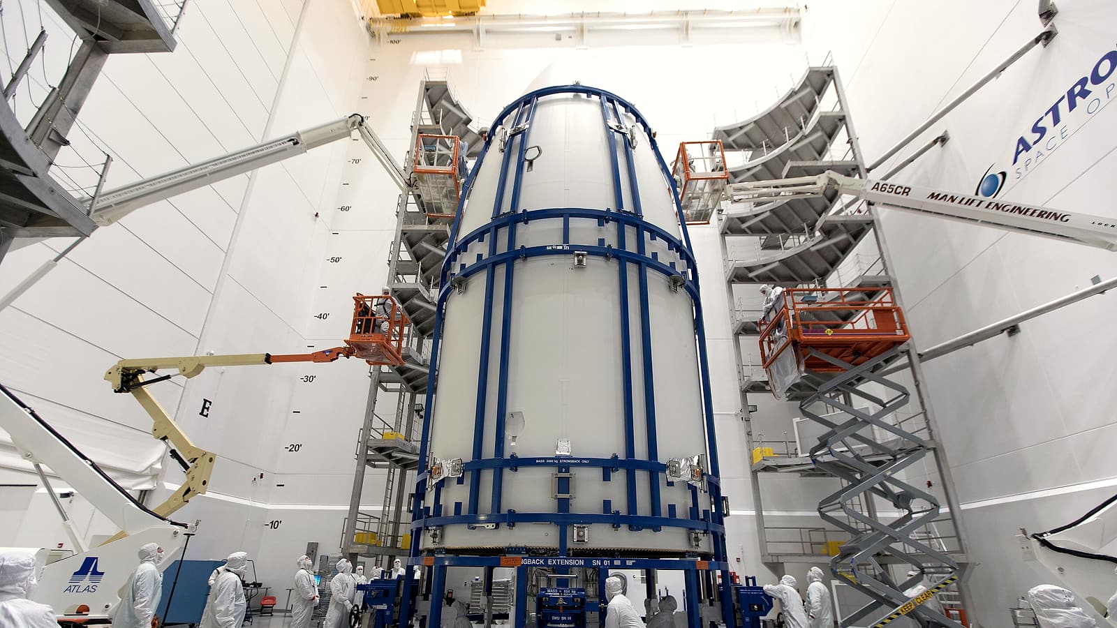 A satellite in a clean room draped in gold foil while two technicians in white coveralls work beside it.