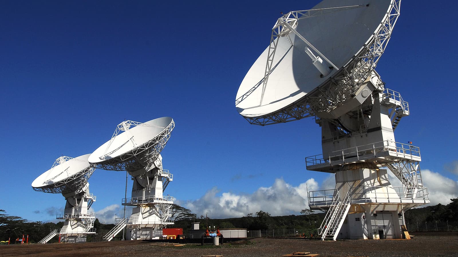 A row of large white parabolic dish antennas on concrete pads pointing at a clear sky above dry grassland.